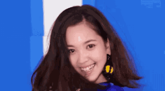 a woman wearing a blue shirt and yellow earrings smiles with her hair blowing in the wind