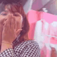 a woman in a plaid shirt is covering her mouth with her hands in front of a pink wall .
