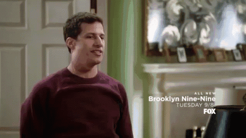 a man in a maroon sweater is sitting in front of a fireplace with a brooklyn nine-nine advertisement behind him