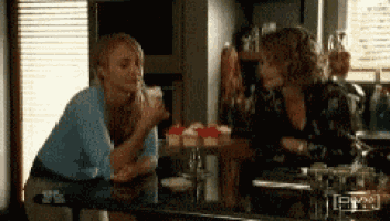 two women are sitting at a table with cupcakes on it and one is kissing the other on the cheek .