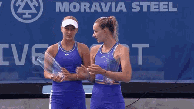 two female tennis players holding trophies in front of a banner that says moravia steel