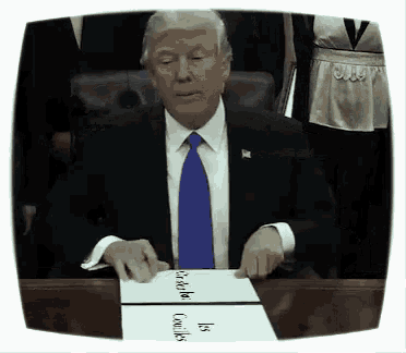 a man in a suit and tie is sitting at a desk with a piece of paper that says ' donald trump is '