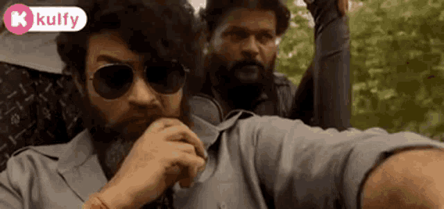 a man with a beard wearing sunglasses is standing next to another man in a car .