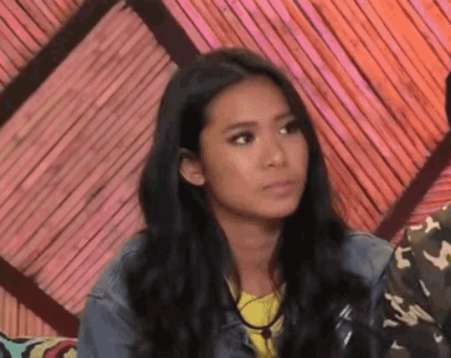 a woman with long black hair is sitting in front of a wooden wall and making a funny face .