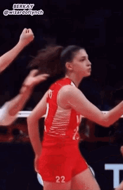 a woman in a red tank top and shorts with the number 22 on her shorts is dancing on a volleyball court .