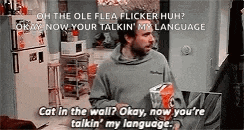 a man is standing in a kitchen holding a box of cat food and talking to another man .