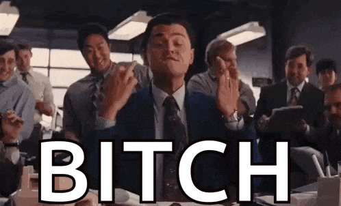 a man in a suit and tie is giving the middle finger in front of a group of people and the word bitch is on the screen .