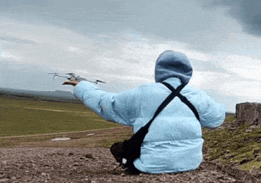 a person in a blue jacket is sitting on a rock watching a drone take off