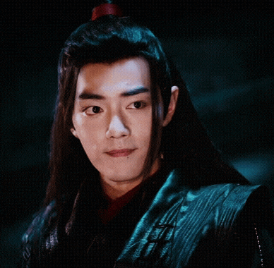 a young man with long black hair is wearing a black jacket and looking at the camera .