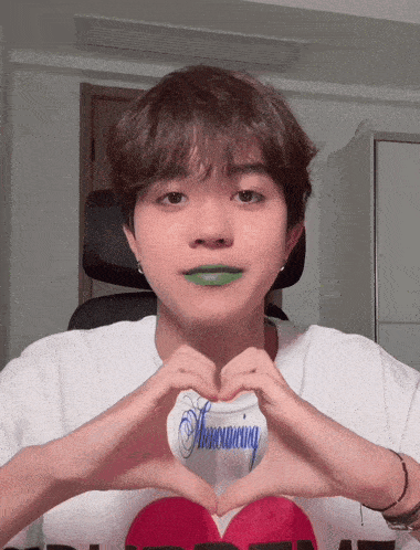 a young man with green lipstick on his lips makes a heart shape with his hands in front of his face