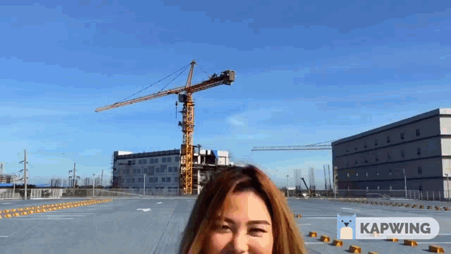 a woman is standing in front of a construction crane and a sign that says kapwing on it