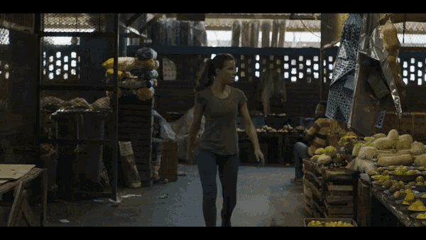a woman in a grey shirt is walking through a fruit stand