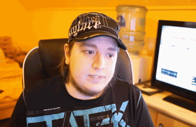 a man wearing a hat and a shirt that says tater sits in front of a computer monitor