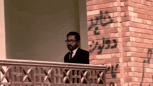 a man in a suit and tie stands on a balcony with a brick wall behind him