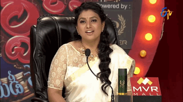 a woman sitting in front of a mvr mall sign speaking into a microphone