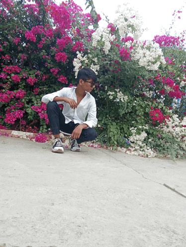 a young man sits on the ground in front of a bush of pink and white flowers