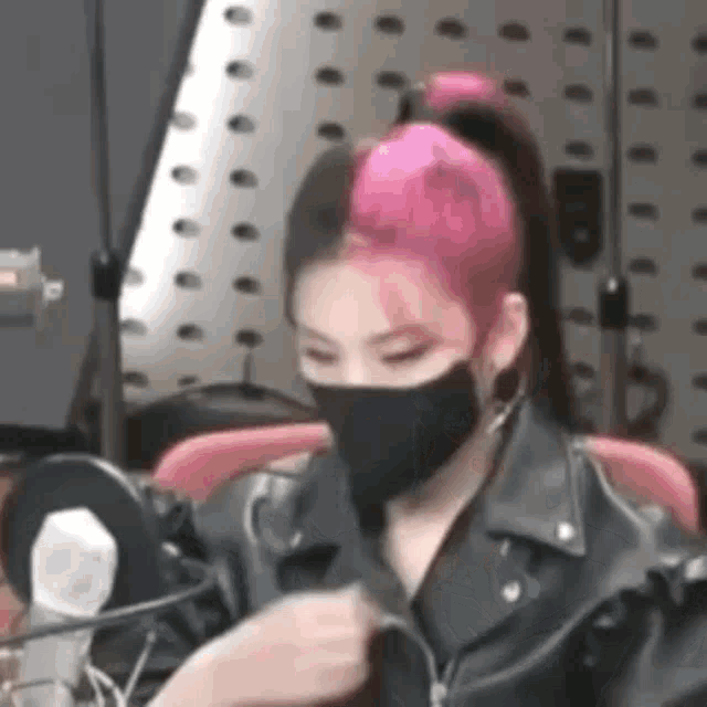a woman with pink hair wearing a black mask and a leather jacket is sitting in front of a microphone .