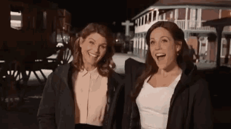 two women are standing next to each other in front of a building and laughing .