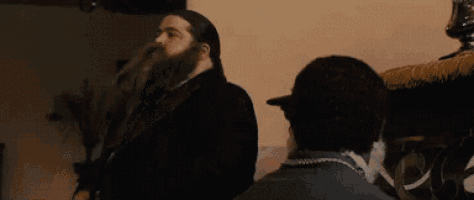 a man with a beard is covering his face with his hands while talking to another man in a room .