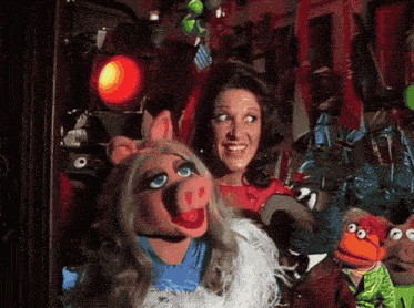 a woman stands next to a pig puppet and another puppet