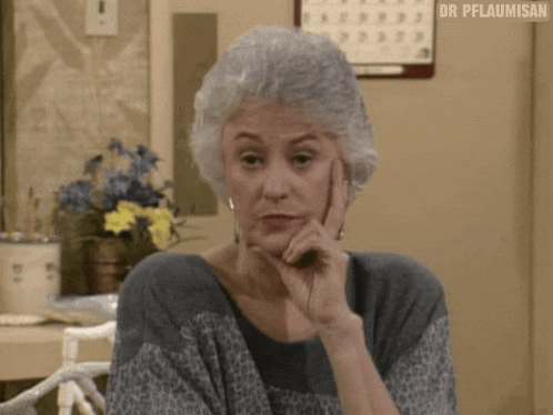 a woman with gray hair is sitting at a table with her hand on her chin and a calendar on the wall behind her .