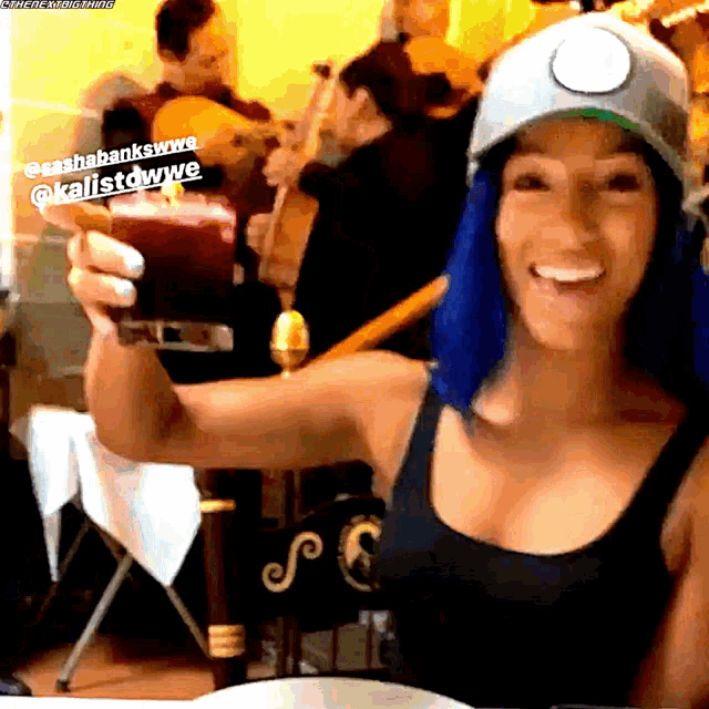 a woman with blue hair wearing a hat holds up a drink
