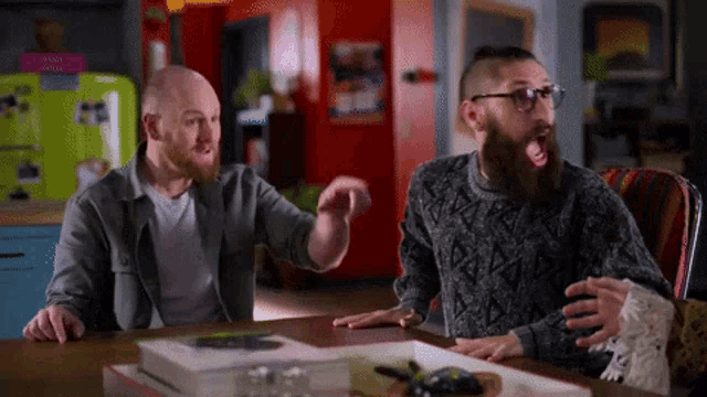 two men are sitting at a table with their mouths open and one is wearing glasses