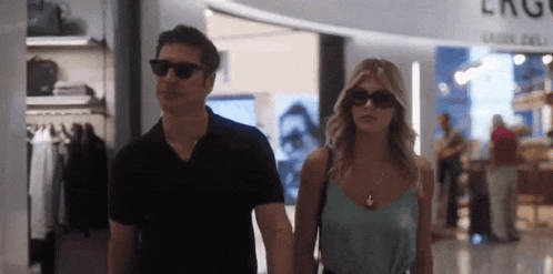 a man and a woman wearing sunglasses are walking in a mall holding hands .