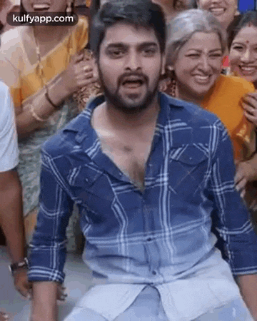 a man in a blue plaid shirt is sitting in front of a crowd of people and making a funny face .