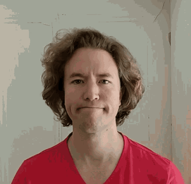 a man with curly hair wearing a red shirt looks at the camera