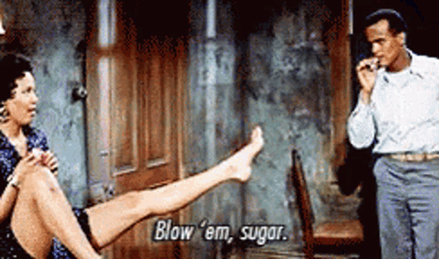 a man is smoking a cigarette while a woman sits on the floor with her legs crossed and says blow em sugar
