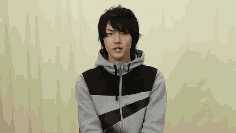 a young man wearing a gray and black hoodie is standing in front of a white curtain .