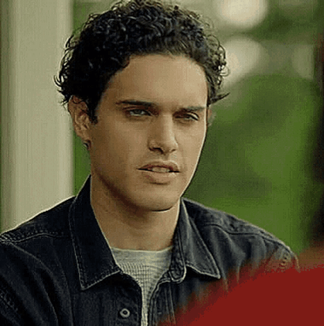 a young man with curly hair is wearing a black denim jacket and a grey shirt .