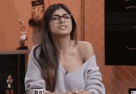 a woman wearing glasses and a sweater is sitting at a table and making a funny face .