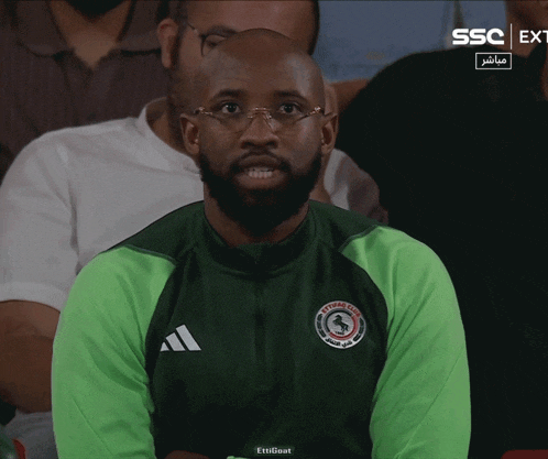 a man wearing glasses and a green adidas jacket is sitting in the audience