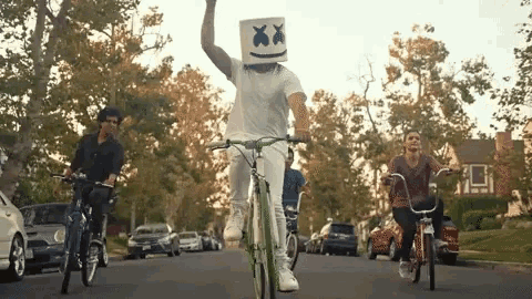 a man wearing a marshmallow head is riding a bike down a street with other people .