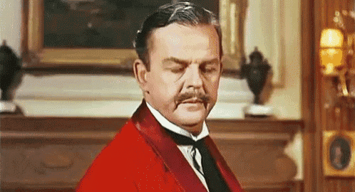 a man with a mustache and a red jacket is standing in front of a fireplace .