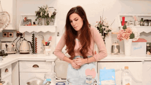 a woman standing in front of a kitchen counter with a blender and flowers on the shelf