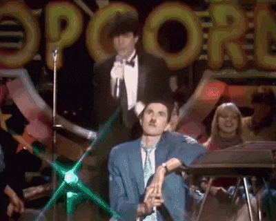 a man in a tuxedo is sitting in front of a microphone on a stage in front of a crowd .