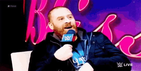 a man with a beard is talking into a microphone while sitting in a chair .