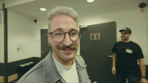 a man with glasses and a mustache is smiling in front of a door that says 2