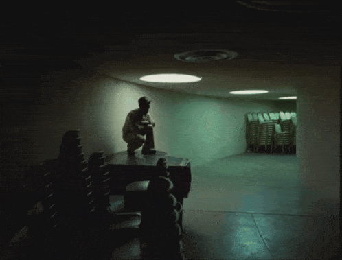 a man sits on a table in a dark room with stacks of chairs in the background