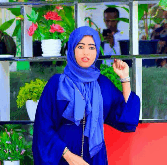 a woman in a blue hijab is standing in front of a shelf with potted plants .