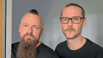 two men with beards and mustaches are standing next to each other in front of a window .