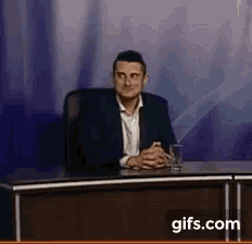a man in a suit is sitting at a desk with his hands folded and a glass of water .