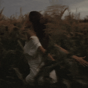 a woman in a white shirt is standing in a field of tall grass with her hair blowing in the wind .