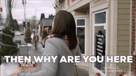 a woman is standing in front of a building with the words `` then why are you here '' written on it .