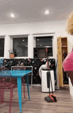 a woman in a pink skirt is standing in a living room with a table and chairs .