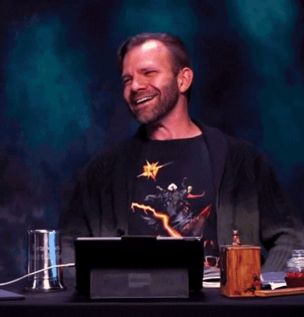 a man wearing a black shirt with a lightning bolt on it is sitting at a desk