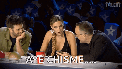 three people are sitting at a table and one of them is holding a cell phone and says aye chisme